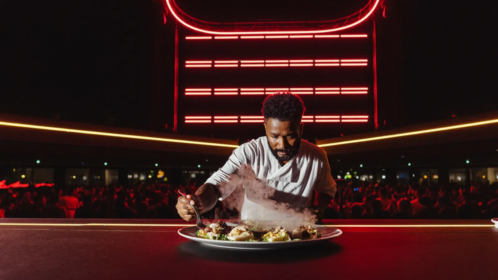 A chef plating a dish under scarlet stage lighting with a crowd behind.